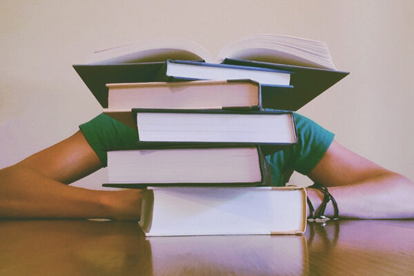 Finals Week: How To Use Caffeine, Sleep And Snacks. A Person Who Is Studying Has Their Head On The Table Behind A Pile Of Books.