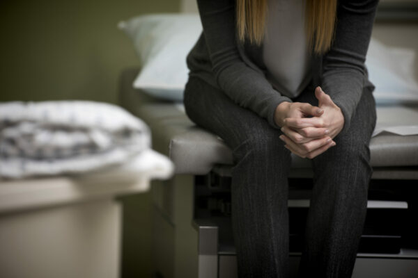 Retrovirus_Autoimmune Disease_Women's Health_A Women Is Sitting On An Exam Table With Her Hangs Resting On Her Legs