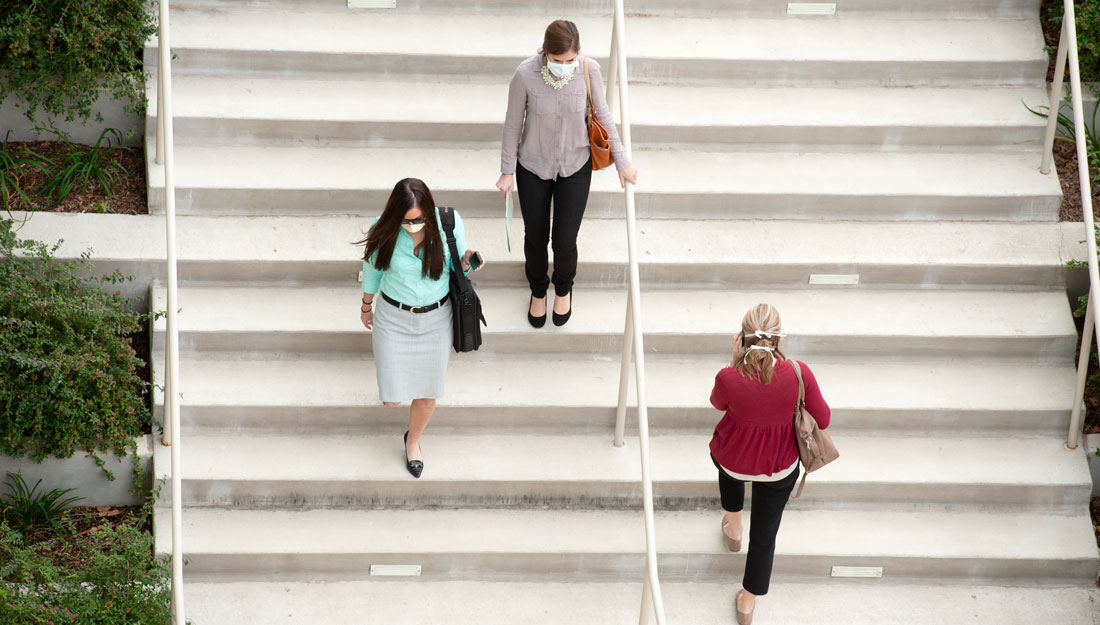 Three women walk on staircase, each wearing a surgical mask.