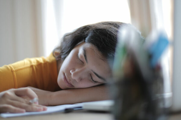 Woman In Yellow Shirt Sleeps At Desk With Her Head Resting On Her Arms