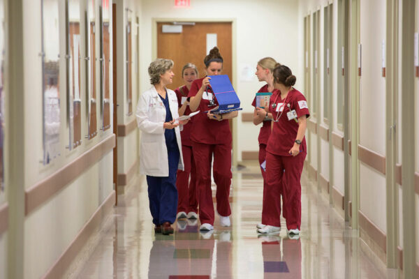 Photo Of Nursing Students In Scrubs Walking Down A Hallway With A Professor In A White Coat