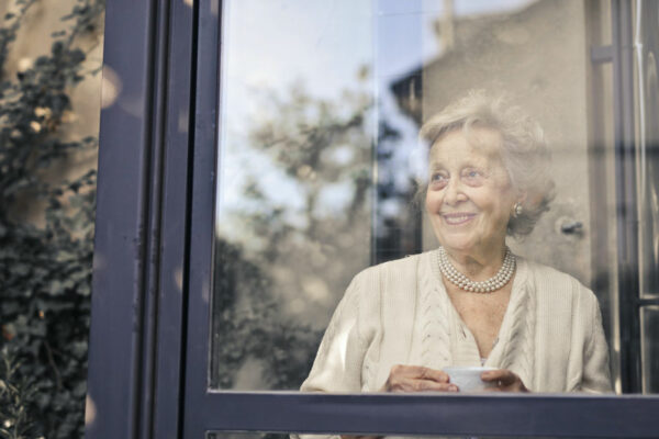 Older Woman Stands In Front Of A Window, Smiling And Holding A Cup Of Tea Or Coffee