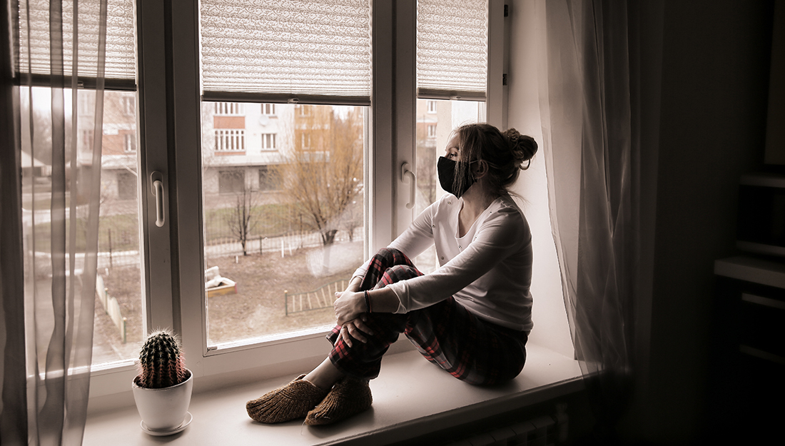 А young woman in a protective mask sits on the window sill. Du