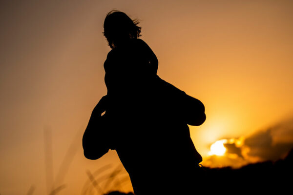 Father Carries Young Daughter On His Shoulders As Sun Sets Behind Them