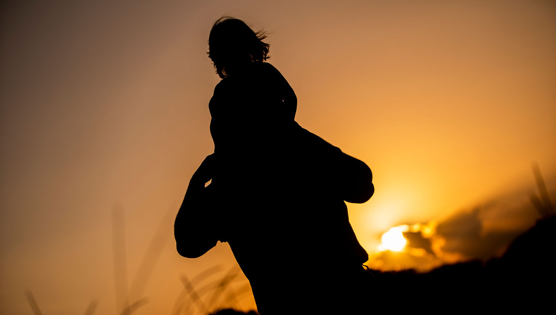 father carries young daughter on his shoulders as sun sets behind them