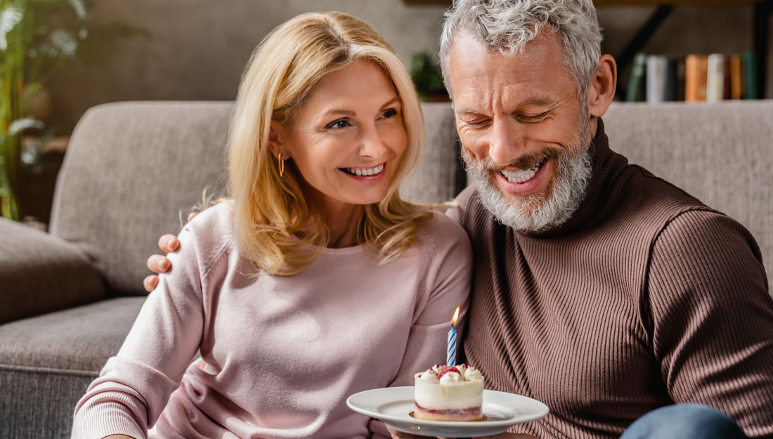 middle aged man sits with woman on couch while holding a cupcake with a candle