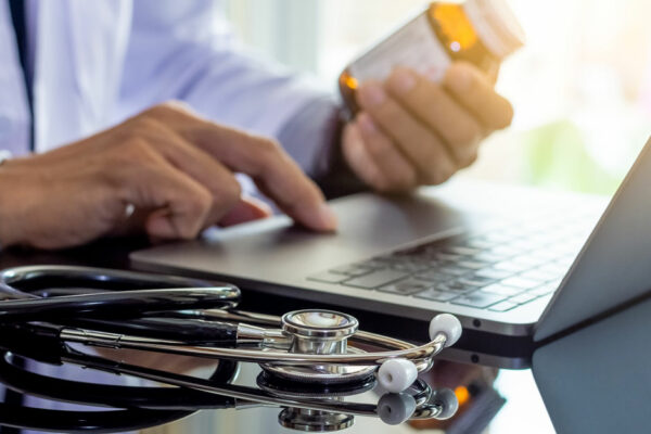 Doctor In White Coat Holding Prescription Bottle While Working On Laptop Computer, Medical Stethoscope On The Desk