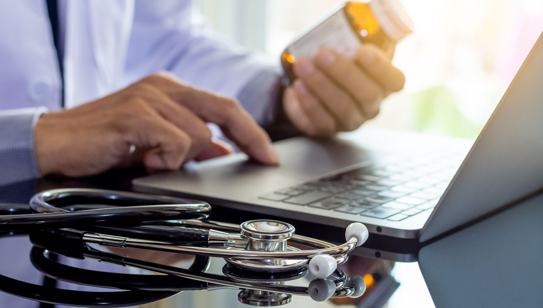 Doctor in white coat holding prescription bottle while working on laptop computer, medical stethoscope on the desk