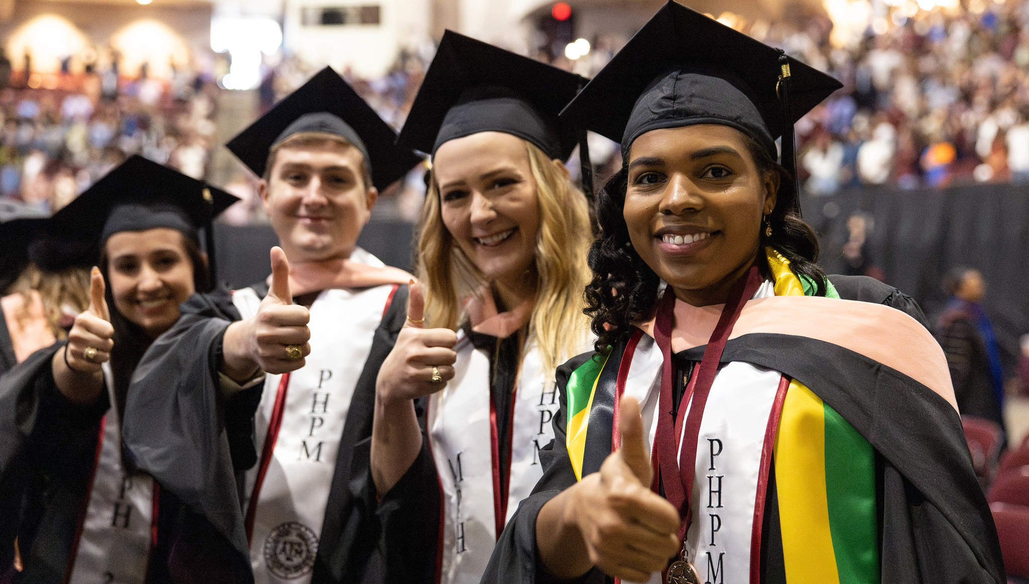 adults wearing graduation regalia holding their thumbs up