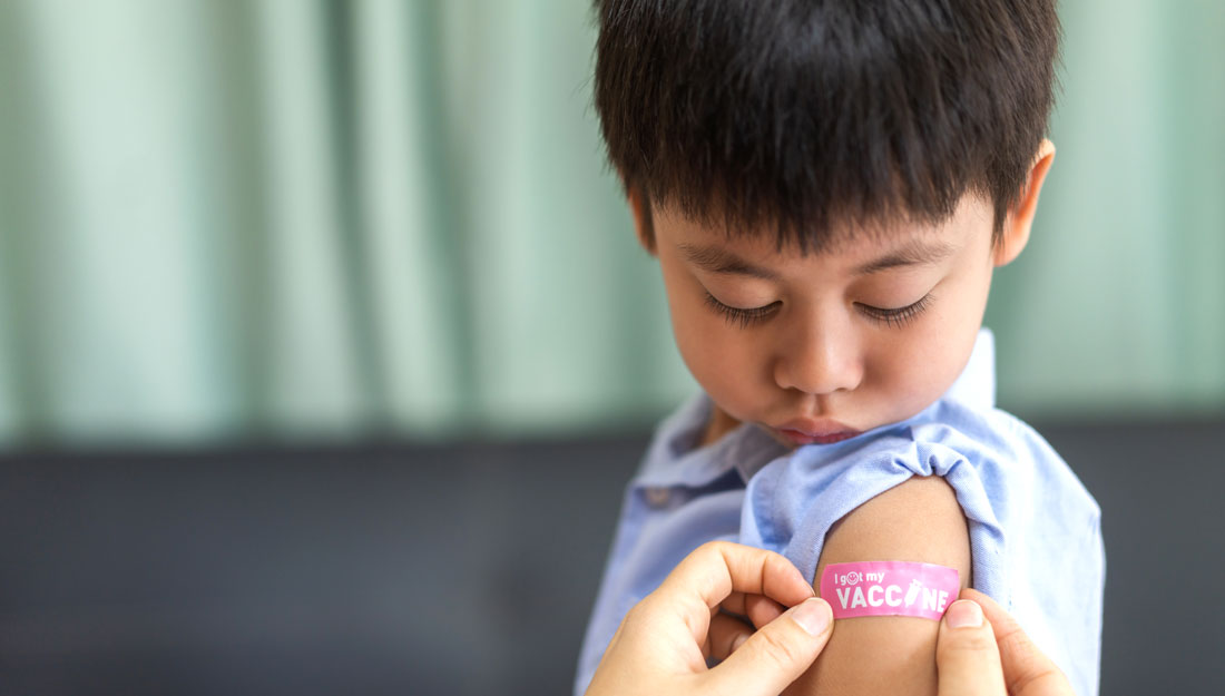 boy looks down at bandaid being placed on his arm after receiving a vaccination