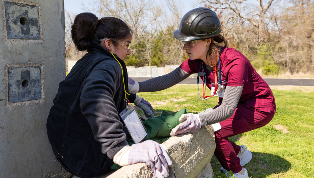 nursing student in maroon scrubs assists another student playing the part of a disaster victim