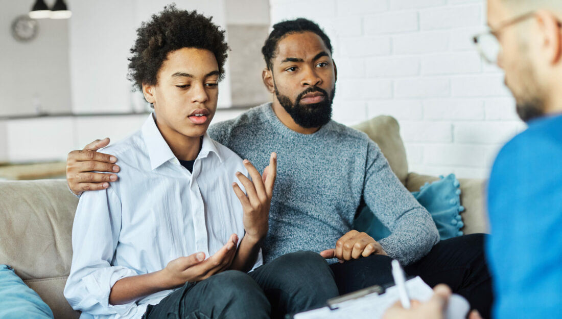 child sits on couch next to father while talking with a professional health care worker