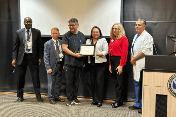 George Martinez Stands On A Stage With A Group, Holding Award