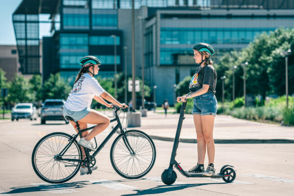 Two Women On Microbility Vehicles—one On A Bicycle And One On A Motorized Scooter—pass Each Other On A Crosswalk In Front Of A University Building