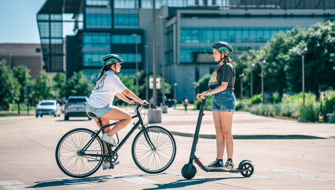 Two women on microbility vehicles—one on a bicycle and one on a motorized scooter—pass each other on a crosswalk in front of a university building