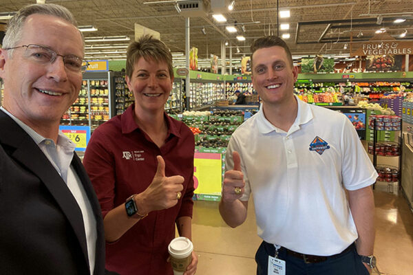 Three Adults Standing In A Grocery Store Pose For A Selfie, Holding Their Thumbs Up In A Gig 'em Sign