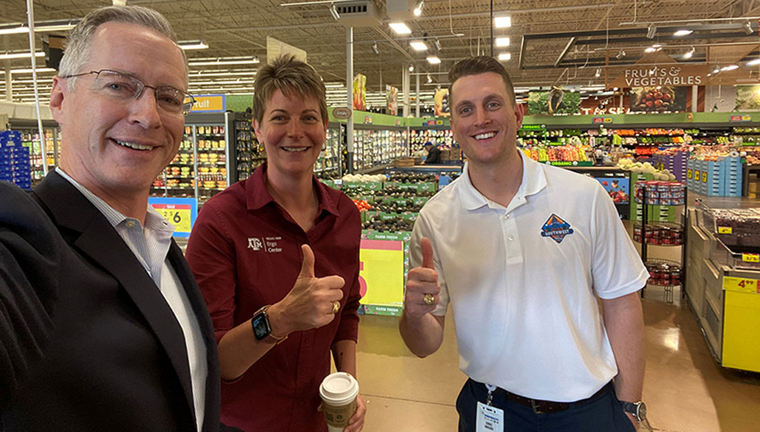 Three adults standing in a grocery store pose for a selfie, holding their thumbs up in a Gig 'em sign