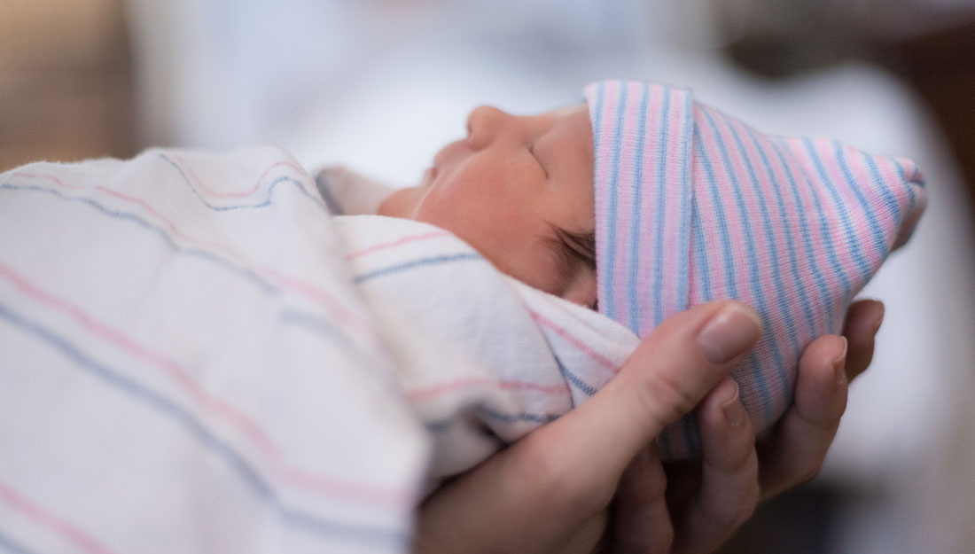 A person’s hands holding a newborn baby wrapped in a striped hospital blanket and wearing a striped beanie