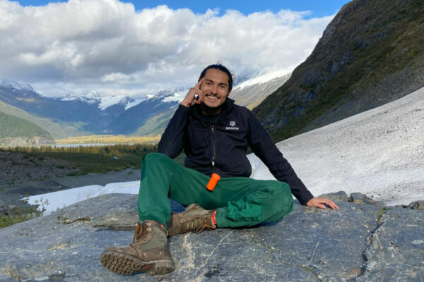 Rodriguez Sits On A Rock Outcrop With Mountains And A Blue Sky In The Background