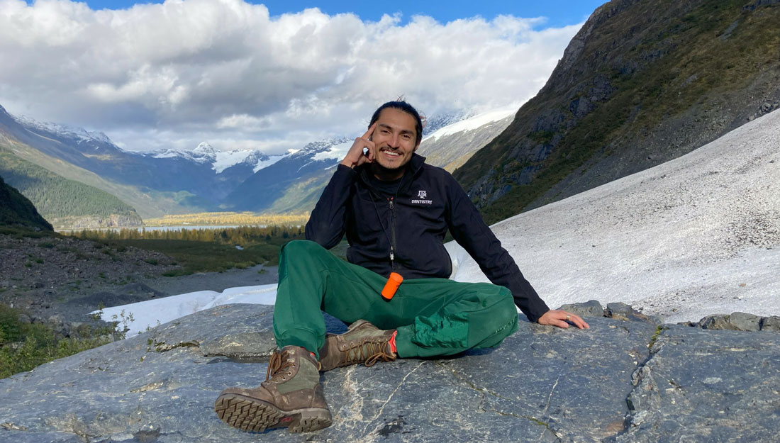 Rodriguez sits on a rock outcrop with mountains and a blue sky in the background