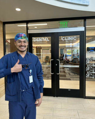 Richard Rodriguez wears dental scrubs and holds up his thumb in a "Gig 'em" sign while standing in front of a dental clinic