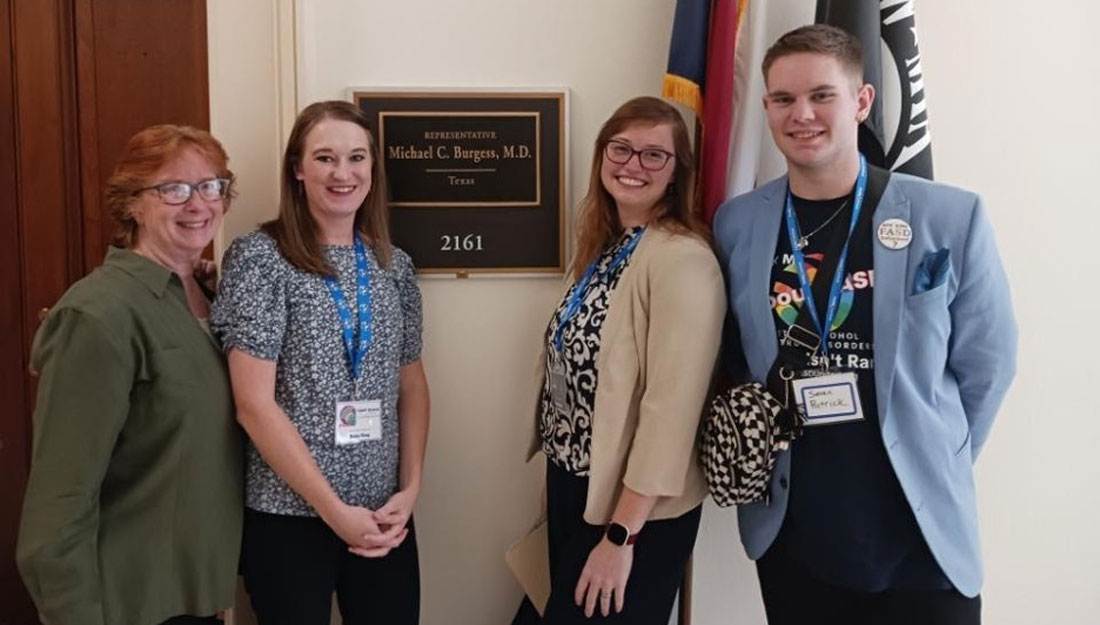 Four people in business attire stand together in front an office sign that says "Representative Michael C. Burgess, MD, Texas"