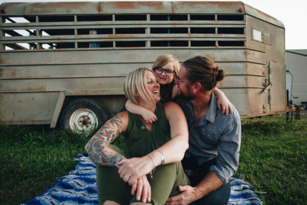 Family Of Three Sits Together On A Blanket In A Field With A Horse Trailer In The Background