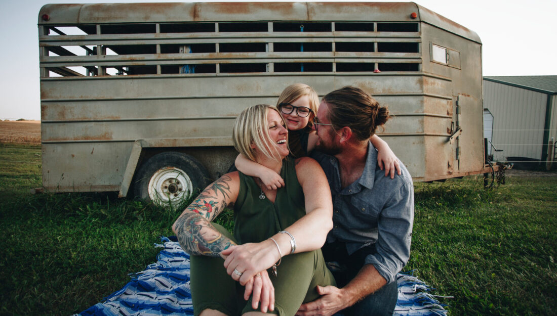 family of three sits together on a blanket in a field with a horse trailer in the background