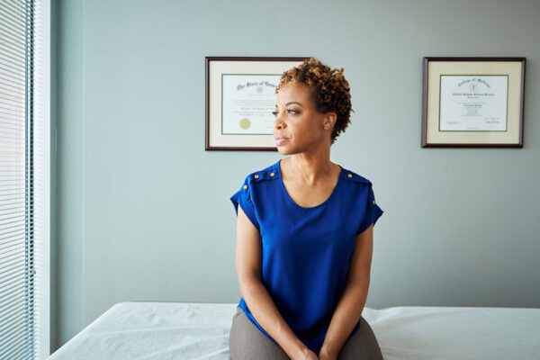 Woman Sits On An Exam Room Table In A Doctor's Office, Looking Out The Window