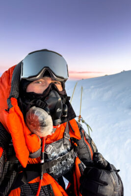 Patrick Monday wears mountain climbing gear and an oxygen mask while standing on the snowy summit of Mount Everest with the sunrise behind him