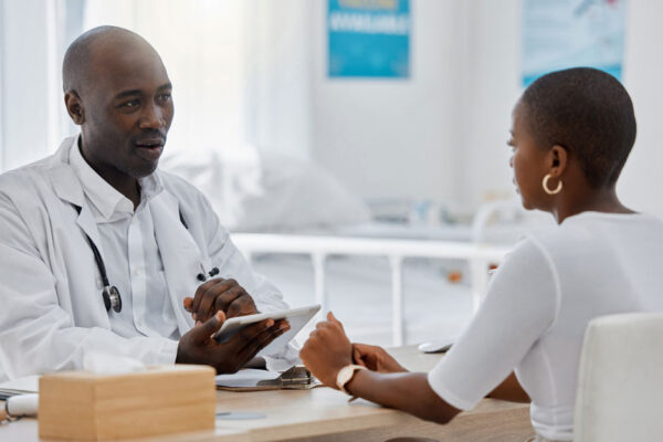 Physician And Patient Sit Across From Each Other At A Desk In A Clinic Room, Physician Holding A Tablet While Speaking