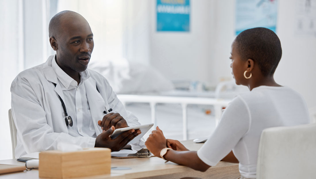 physician and patient sit across from each other at a desk in a clinic room, physician holding a tablet while speaking