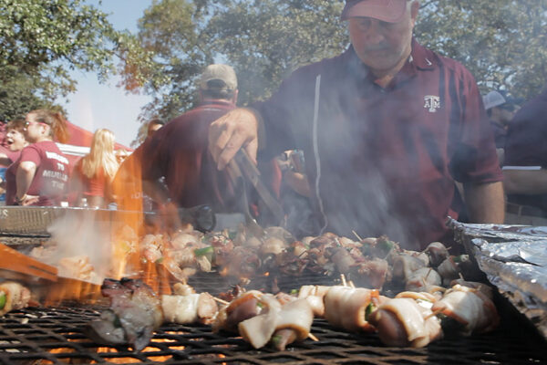 Man Stands Over Grill Flipping Skewer With Tongs, Demonstrating Tailgate Safety