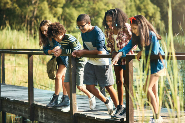 A A Group Of Teenagers Stand On A Bridge In Nature, One Girl Pointing To Something They All Look At