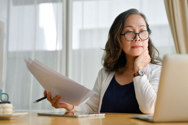 Middle Aged Woman Sitting At Kitchen Table Holds Paperwork And Pen In One Hand While Looking At A Laptop Computer Screen