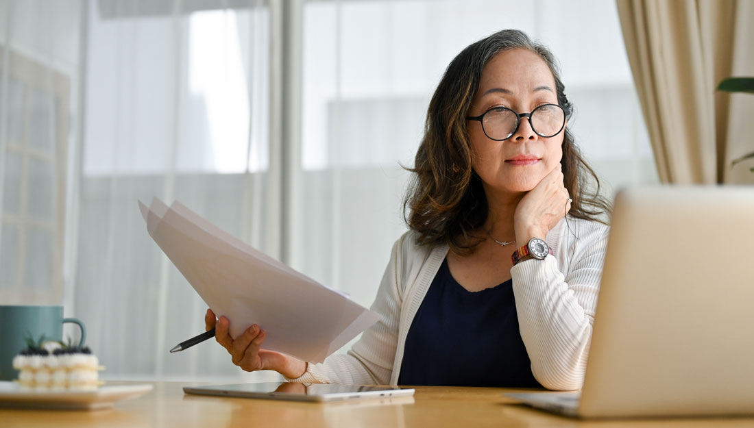 middle aged woman sitting at kitchen table holds paperwork and pen in one hand while looking at a laptop computer screen