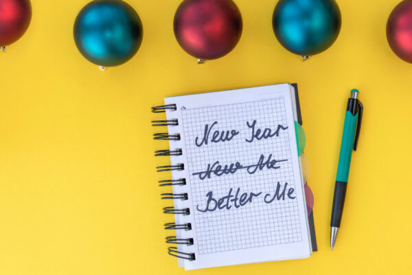 Yellow Backgruond With Blue And Red Christmas Bobbles And A Notepad That Reads "New Year, New Me" With "New Me" Crossed Out And "Better Me" Written Instead.