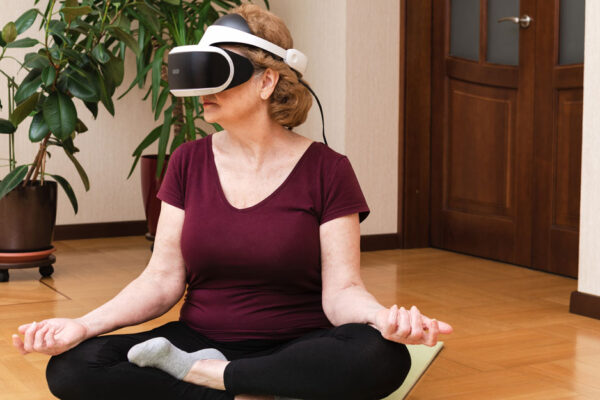 Senior Woman Sitting On Yoga Mat Meditating In VR Glasses At Home. Virtual Reality Meditation