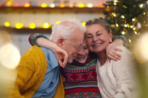 Grandchild Embraces Grandparents In Front Of A Christmas Tree