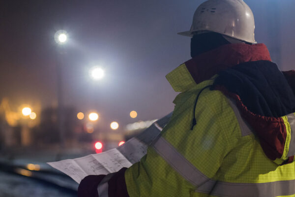 Back View Of Young Construction Worker In Helmet At Night Reading Construction Drawings