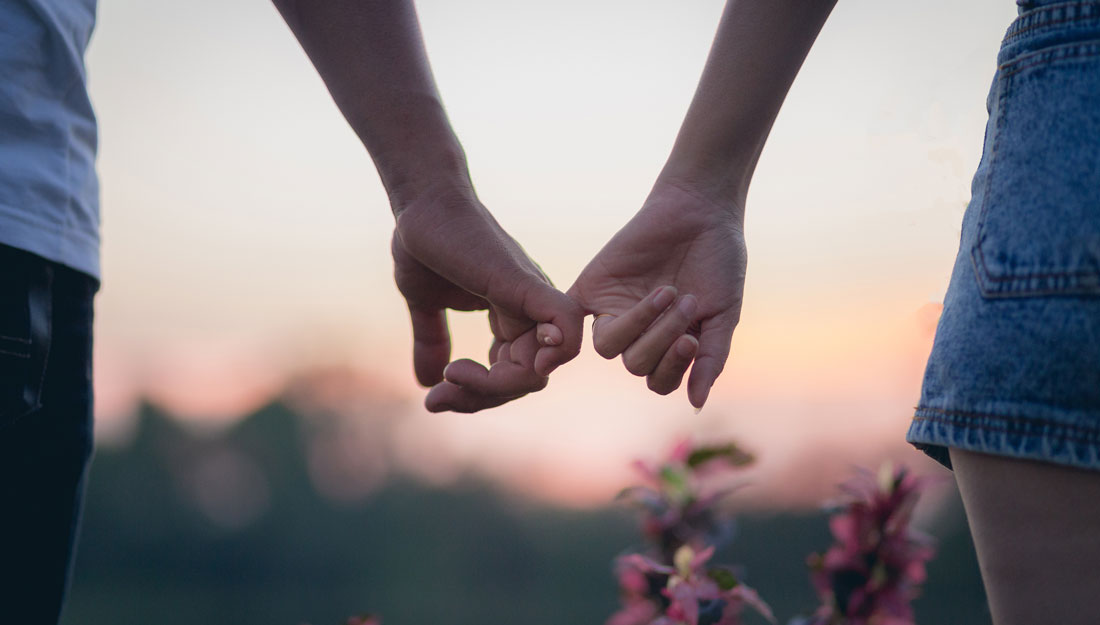 couple locks pinkies while walking through a meadow