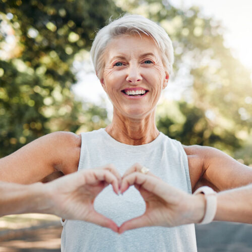 Smiling Older Woman Exercising In Nature Holds Hands Together In A Heart Shape In Front Of Her Chest