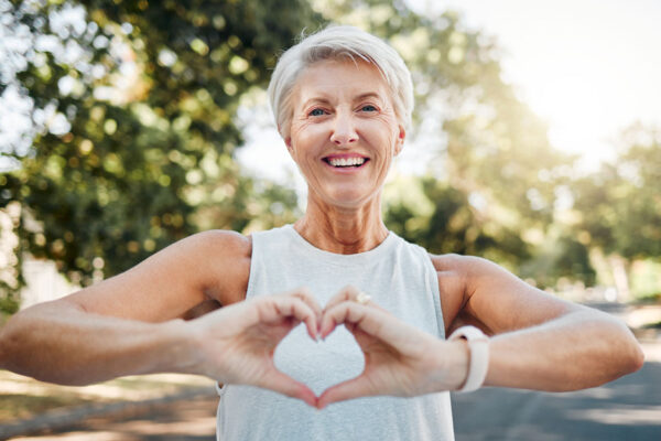 Smiling Older Woman Exercising In Nature Holds Hands Together In A Heart Shape In Front Of Her Chest