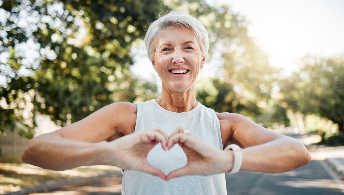 smiling older woman exercising in nature holds hands together in a heart shape in front of her chest