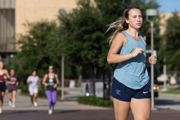 Girl Running In Foreground In Tank Top And Shorts With A&M Campus And Other Runners In The Background