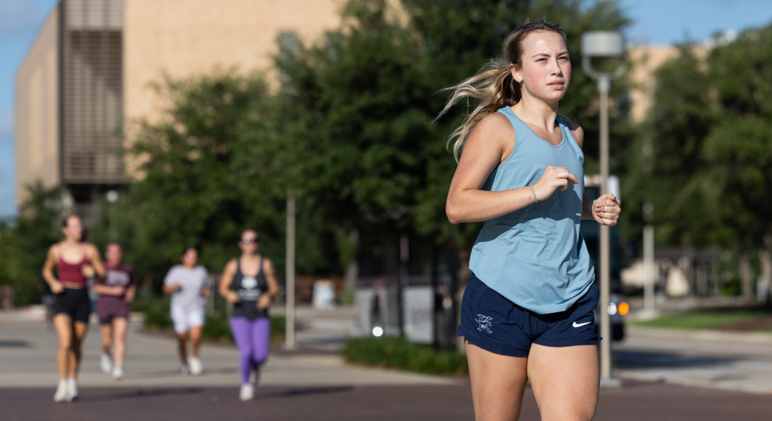 girl running in foreground in tank top and shorts with A&M campus and other runners in the background