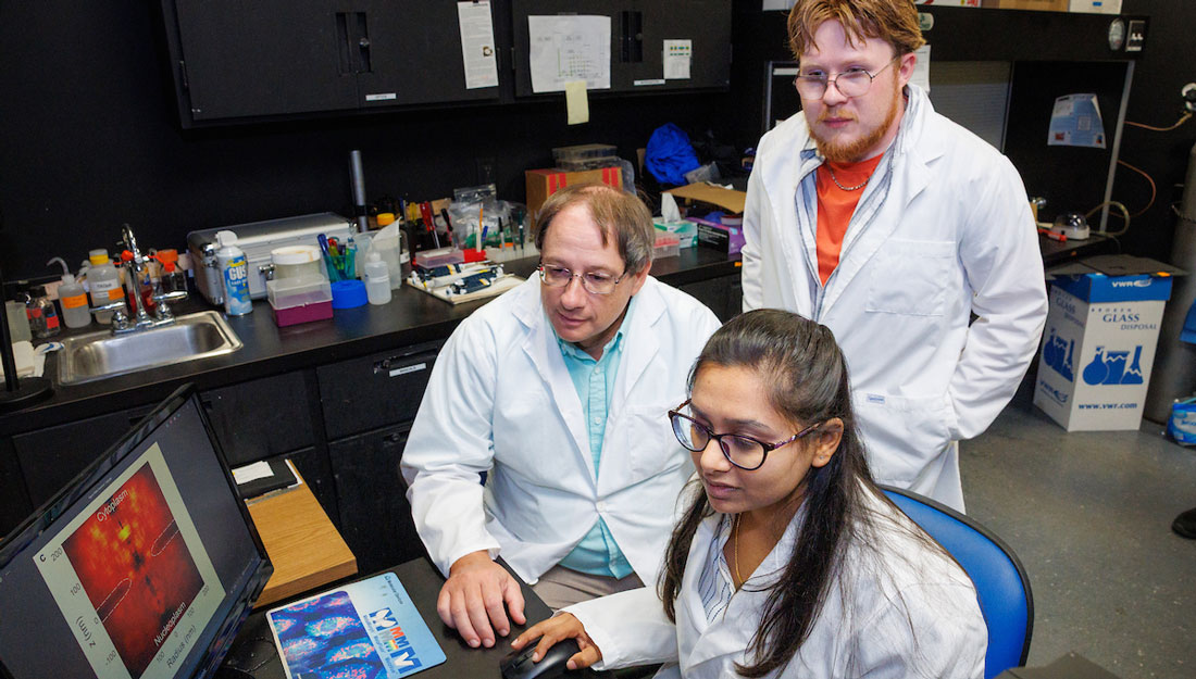 scientists wearing white lab coats look at an image on a computer monitor together