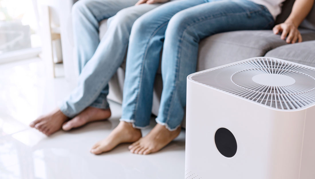 Close up of white air purifier on the floor in living room with a young couple relaxing on a sofa in the background