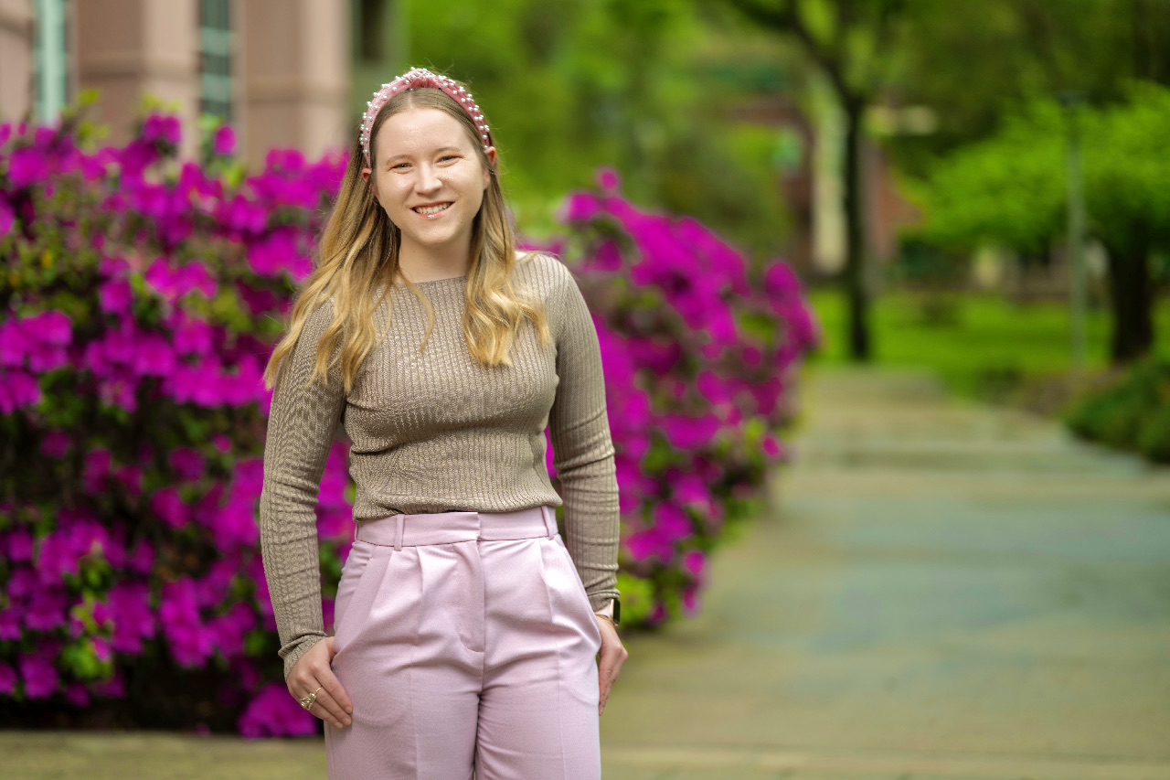 Savannah Kaspar, smiling, stands in a pathway lined with bright pink flowering bushes